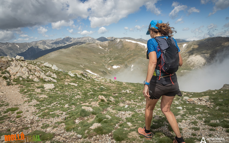Una noia, amb samarreta i gorra blava, d'esquenes caminant per la muntanya amb el nom el logotip de la molló trail a l'esquerra a baix. El logotp és el nom, molló en taronja i trail en verd poma