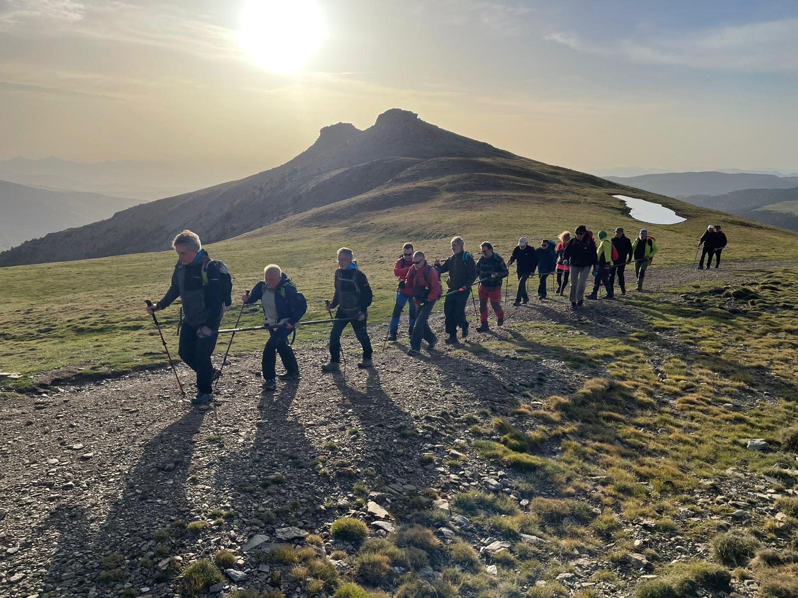 Una filera d'excursionistes caminant amb barres direccionals. Paisatge de muntanya en plena sortida de sol, es veu una parella de cims al fons. 